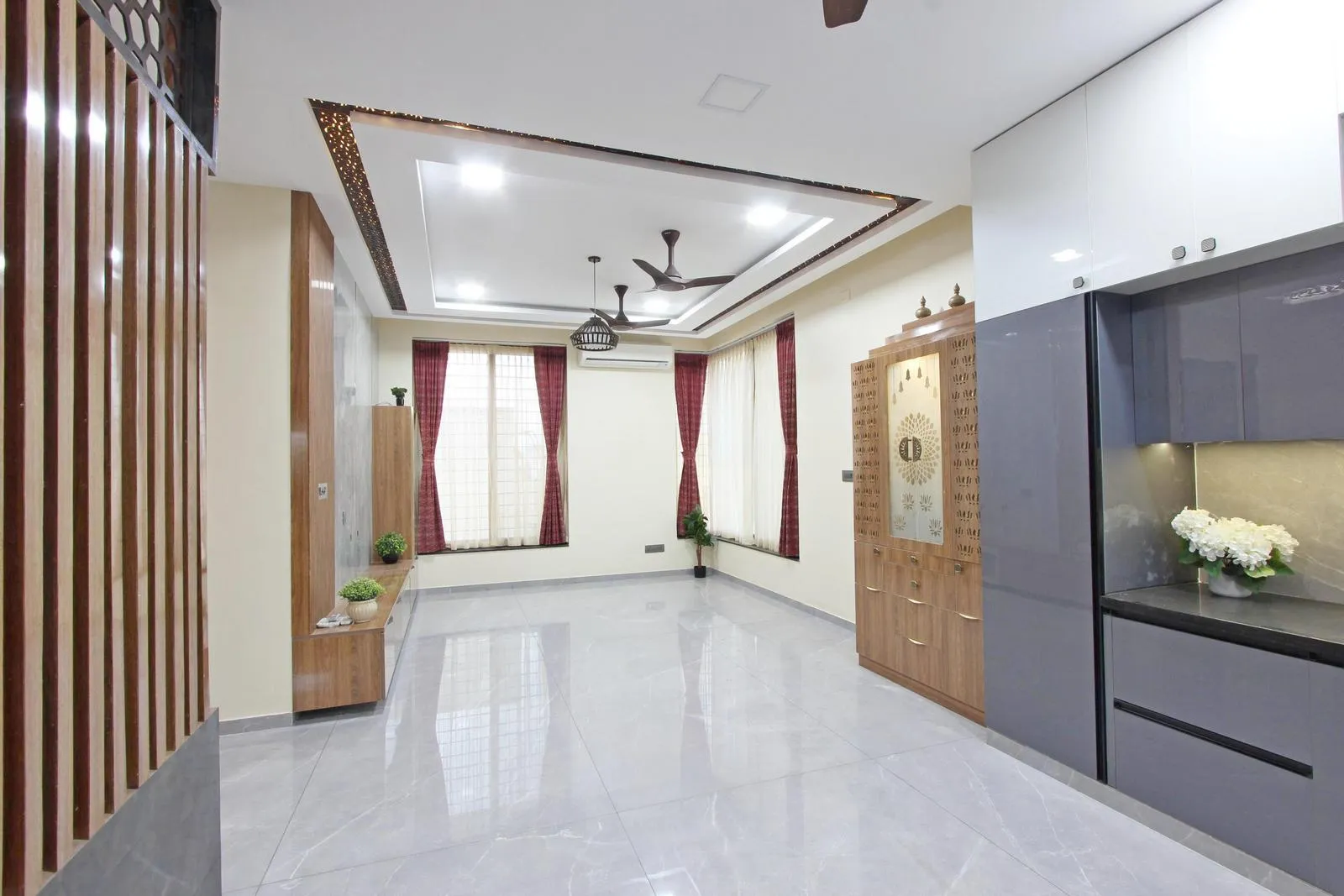 The open-plan living area flows seamlessly into the kitchen and dining zones. Large windows, a teak TV unit, and a wooden puja enclosure add warmth and elegance. Layered lighting, jaali detailing, and floating furniture enhance depth and balance, while natural materials and high-gloss finishes create a modern, harmonious interior.