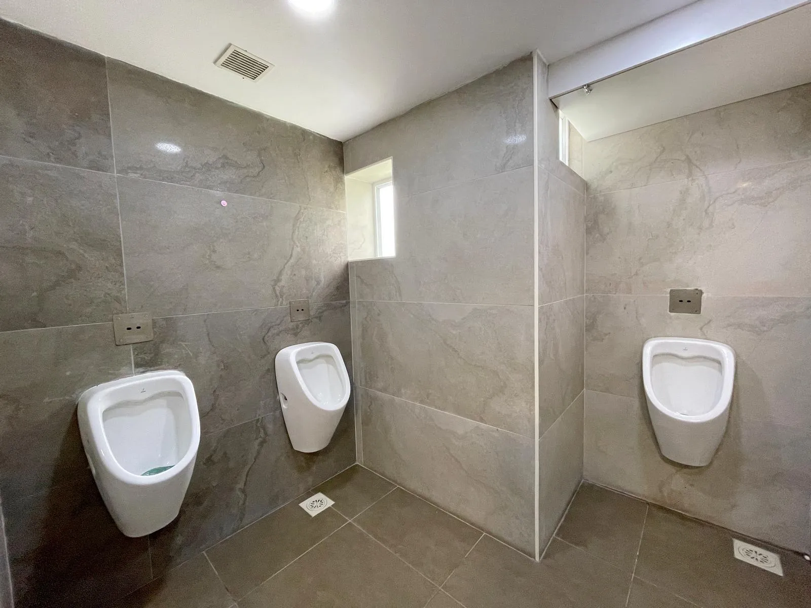 The restroom features wall-mounted urinals, concealed plumbing, and large-format grey marble tiles for a sleek, hygienic finish. Recessed lighting, frosted windows, and strategically placed drains ensure functionality, privacy, and easy maintenance.