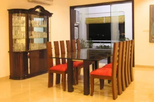 Dining room with a glass-top table surrounded by eight wooden chairs with red cushions, and a dark wooden cabinet displaying glassware in the background, with a view of a balcony at night through sliding glass doors.