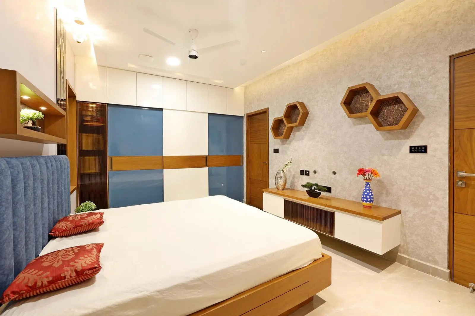 A wardrobe wall as a focal point, and a floating console for openness. Warm wood, white, and blue accents create a balanced, inviting palette. Hexagonal shelves and recessed lighting add depth, functionality, and modern elegance.