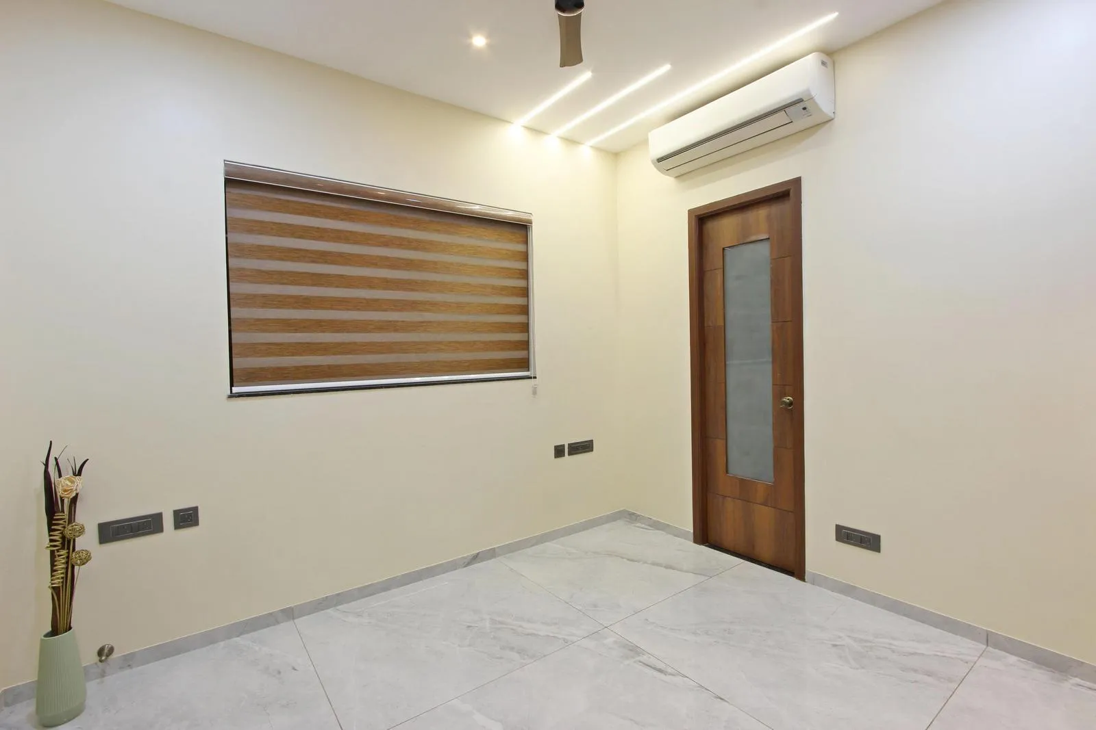 The space features a rectilinear layout with soft beige walls, light grey marble flooring, teak and aluminum accents, and linear LED lighting. Thoughtful placement of doors, windows, and decor ensures natural light, ventilation, and visual balance.