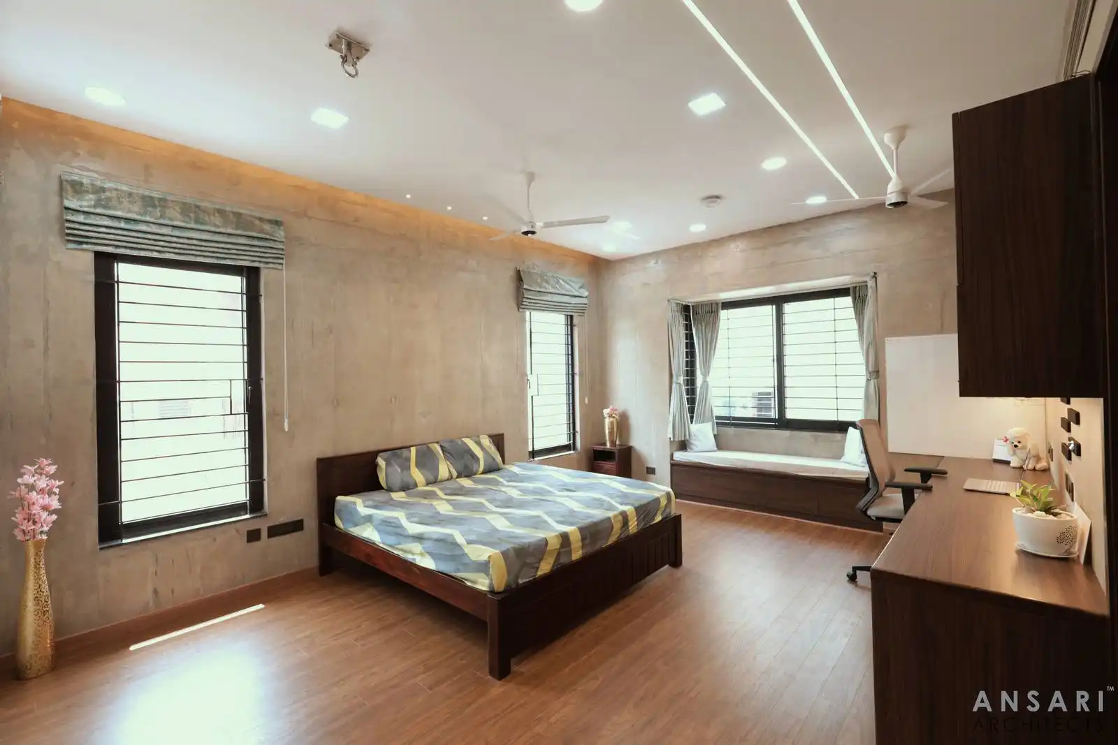 Textured concrete walls and warm wooden floors blend with built-in seating and workspace, enhanced by layered lighting and sleek windows.