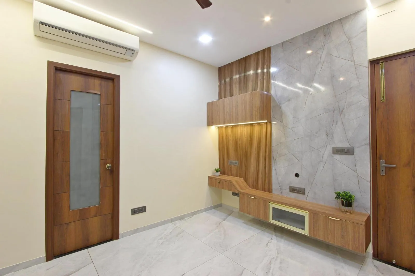 The bedroom centers on a TV wall framed with wood, complemented by a floating console and built-in cabinetry for balanced storage. Light grey flooring, marble-textured walls, and teak finishes create a bright, modern, and inviting space.