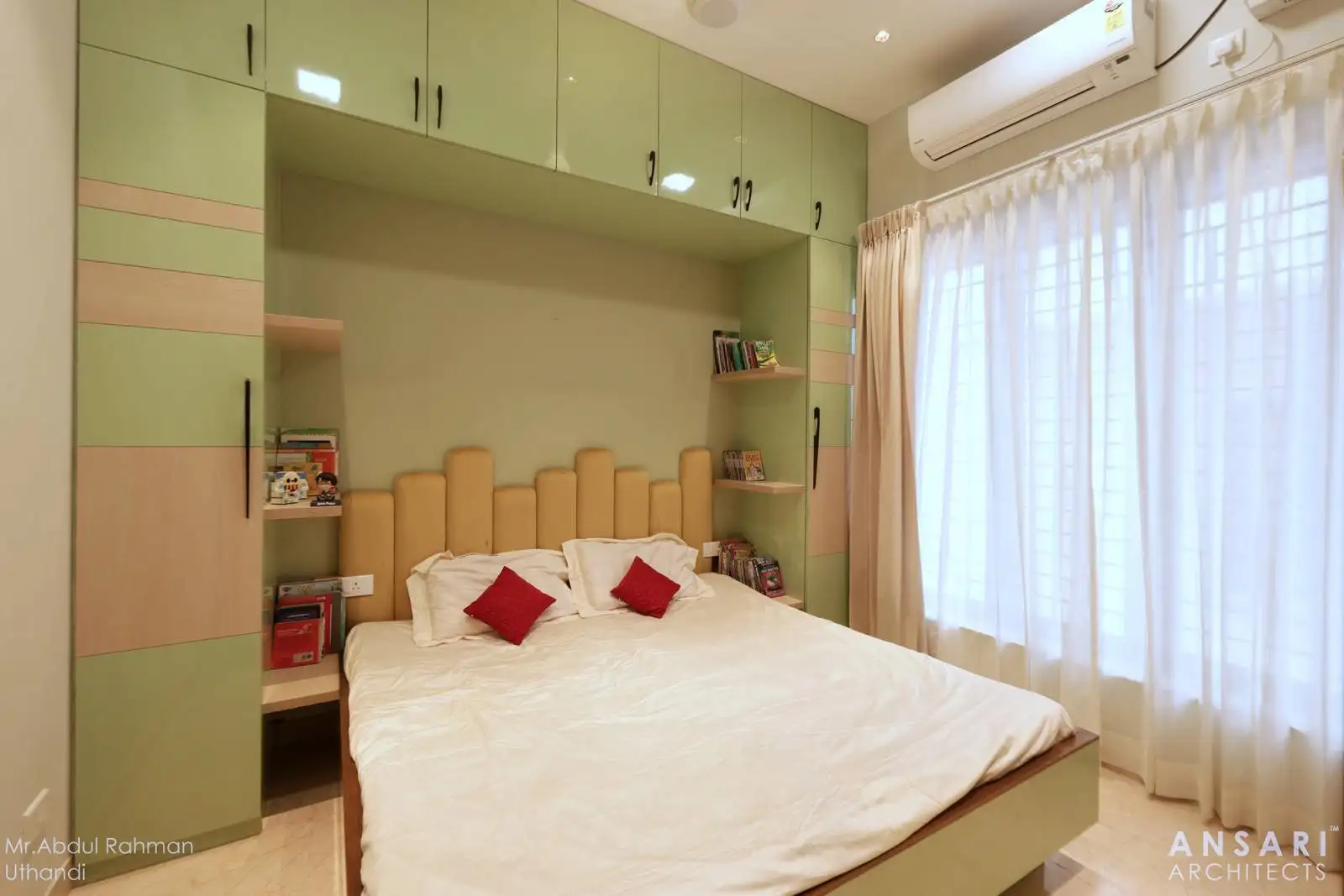 A thoughtfully designed kid’s bedroom featuring a large bed with a padded headboard, accented pillows, and built-in cabinetry with integrated shelving for books and decor. A window with sheer curtains and an air conditioning unit ensures comfort and natural light.
