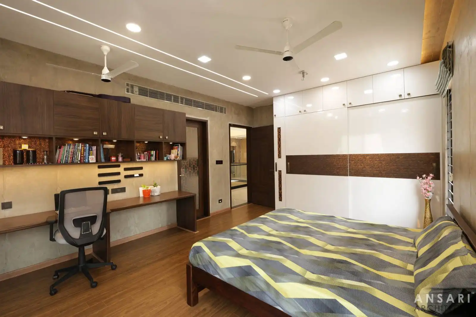 Bedroom with wooden flooring, built-in white wardrobe with brown accent, and a compact work desk with shelves, creating a functional and cozy modern space.