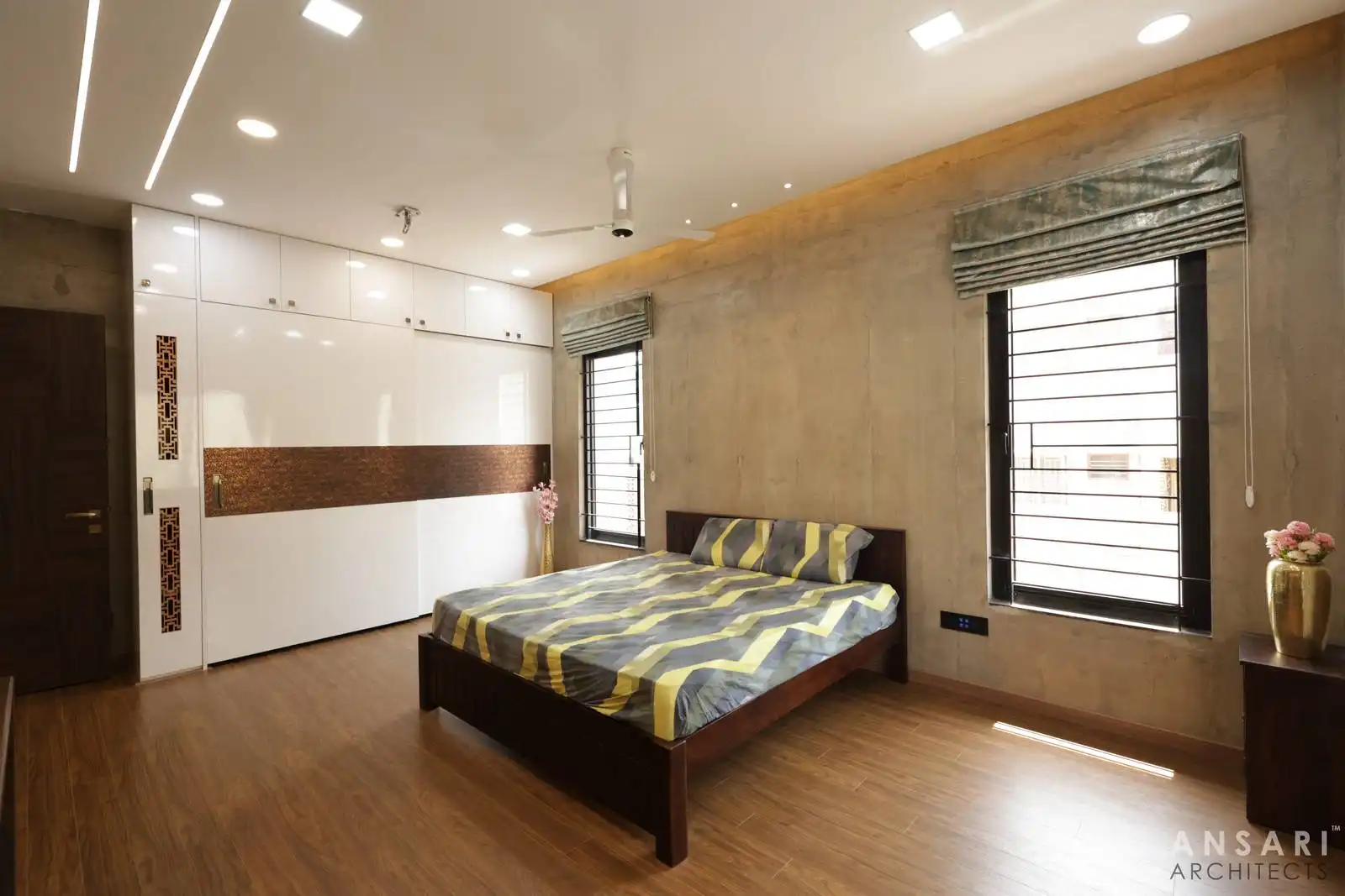 Designed bedroom with polished wooden flooring, textured beige walls, sleek white cabinetry, and large windows with modern blinds, creating a warm, elegant, and functional space.