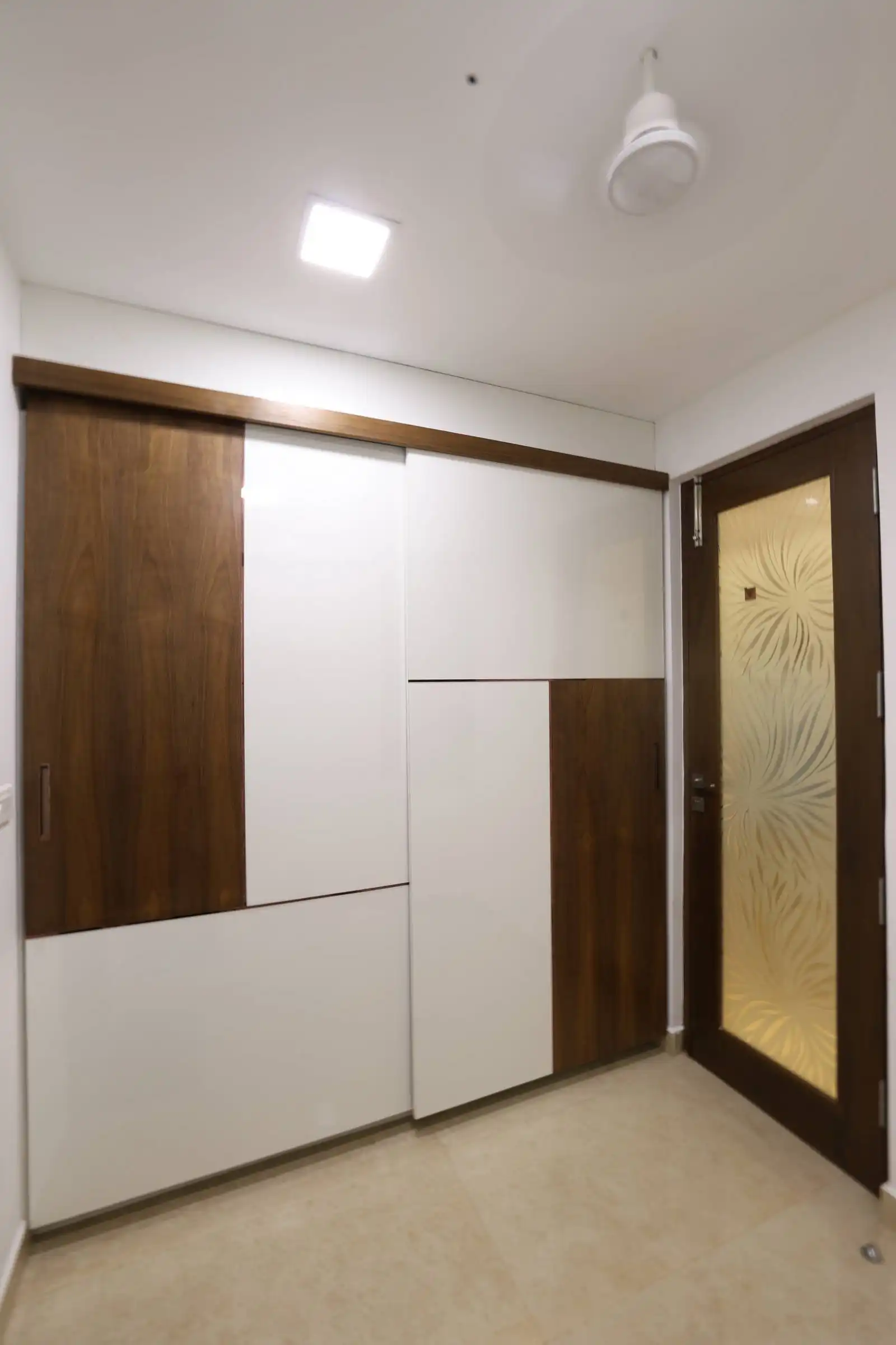 Dressing room features a wooden and white paneled sliding wardrobe alongside a decorative glass door, under a white ceiling with a ceiling fan and recessed lighting, combining functionality and refined design.