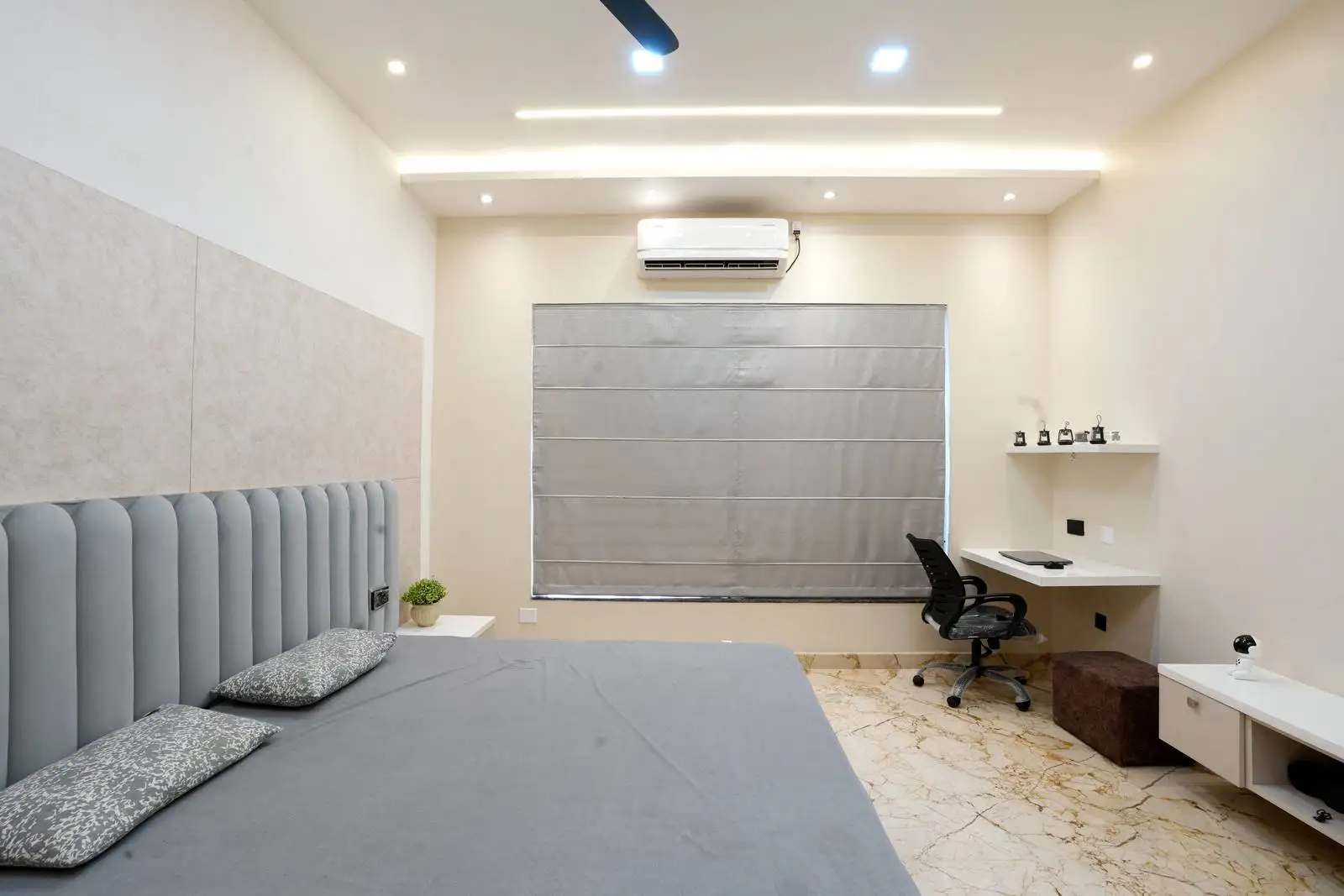 Minimalist bedroom featuring a padded headboard, integrated workspace, and a large window with gray Roman shades for natural light and privacy.