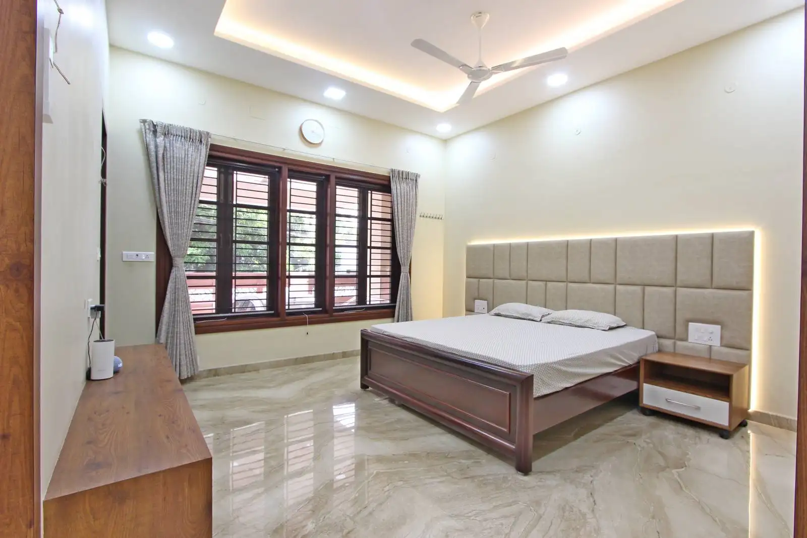 A serene, light-filled bedroom designed with a central layout, rich wood accents, backlit upholstered headboard, and polished marble flooring—balancing luxury, comfort, and functionality.