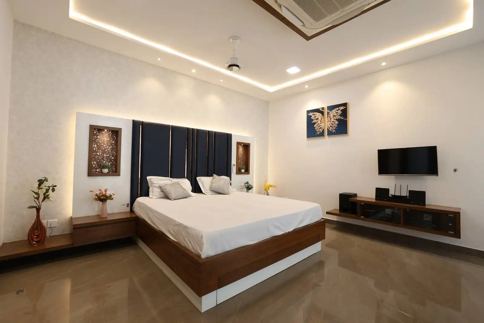 Glossy neutral tiles, warm wood and white finishes, and a deep blue padded accent wall with recessed niches create a spacious, modern, and inviting bedroom.