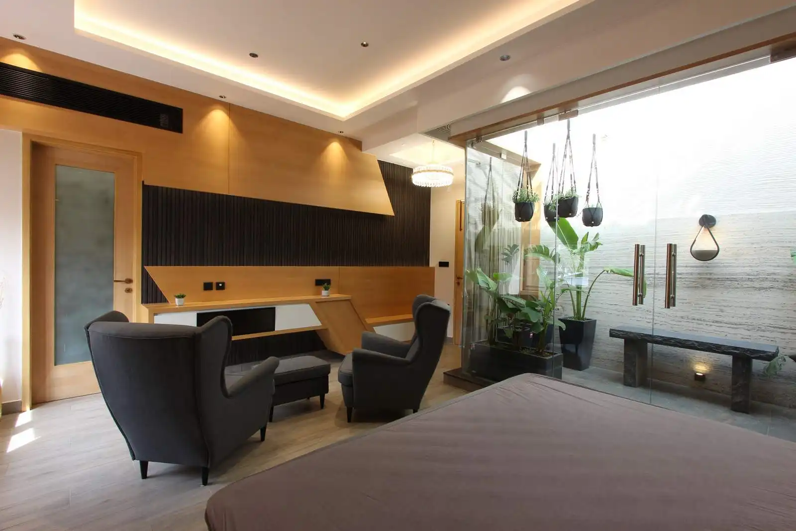 Modern living room with gray armchairs, a wooden desk against a black and wood-paneled wall, large glass doors opening to an outdoor area with hanging plants and a stone bench, and part of a bed with a brown cover in the foreground.