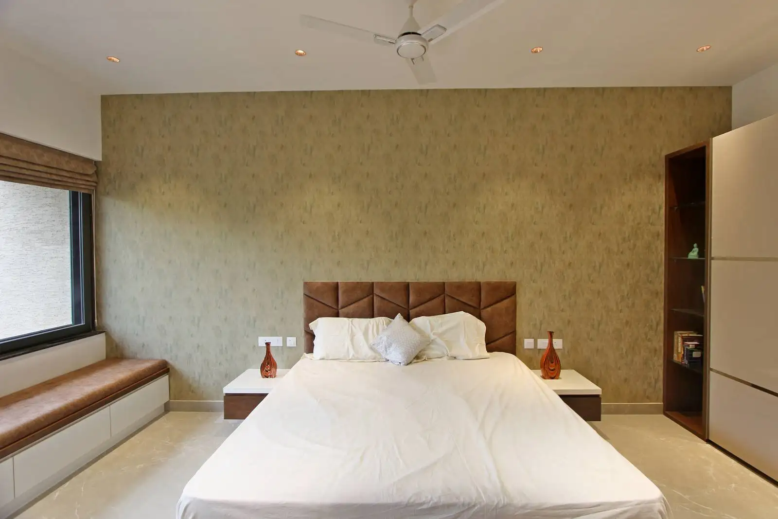The bedroom showcases a textured feature wall behind the bed, adding depth and warmth, complemented by a large window that fills the space with natural light and offers a serene seating area.