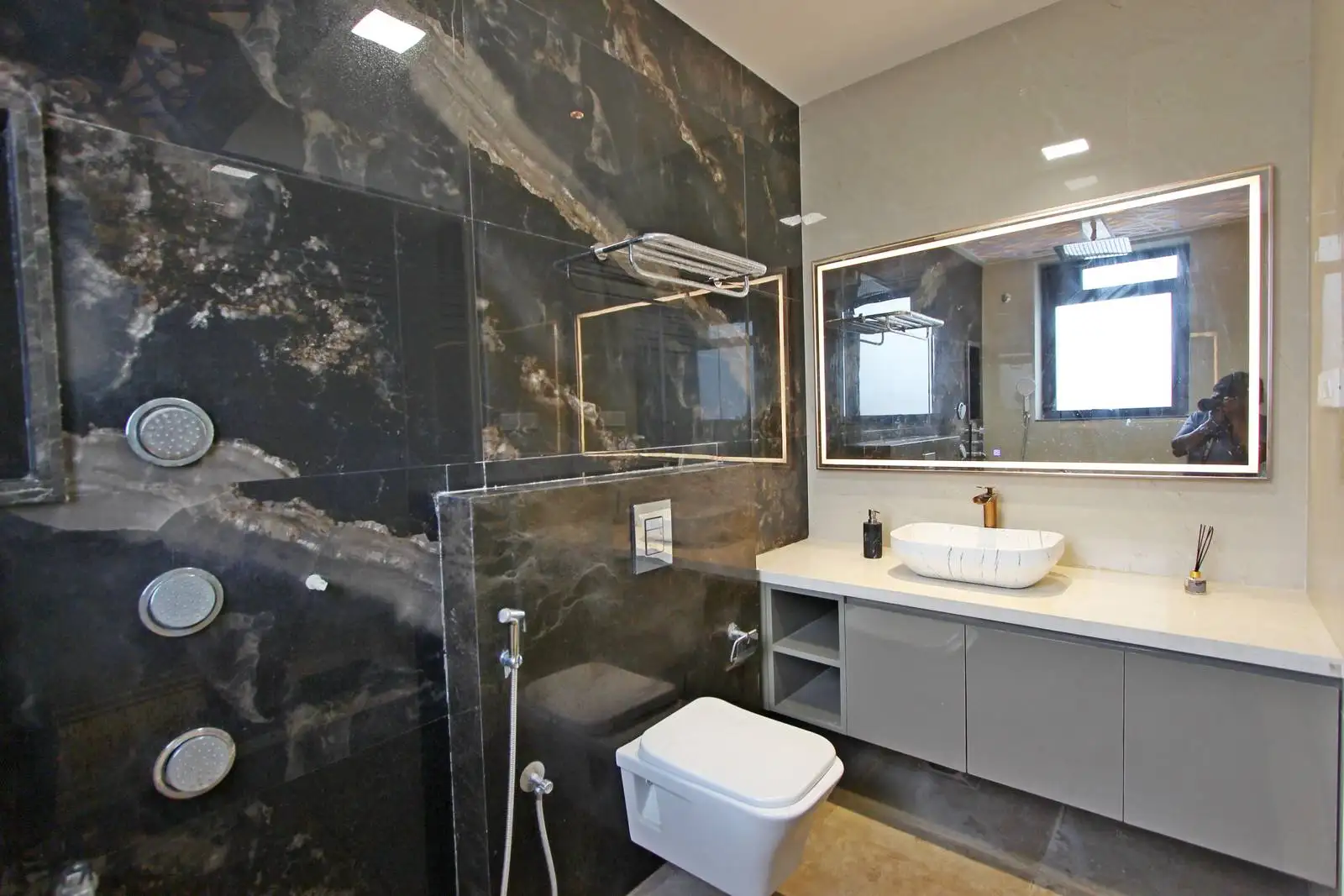A bathroom with dark marble walls, a floating gray vanity with white vessel sink, a floating white toilet, and a large illuminated mirror that reflects the space, creating a sleek and modern aesthetic.