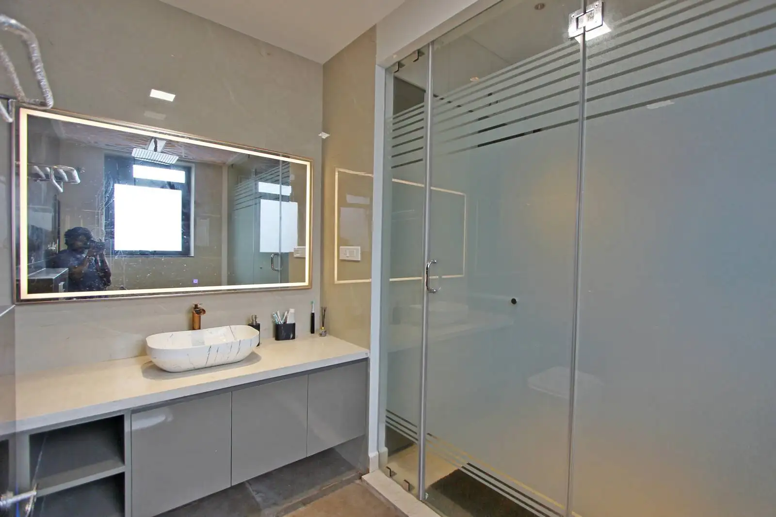 A bathroom with a white marble vessel sink, illuminated mirror, and frosted glass shower with sleek metal accents, blending elegance and functionality.