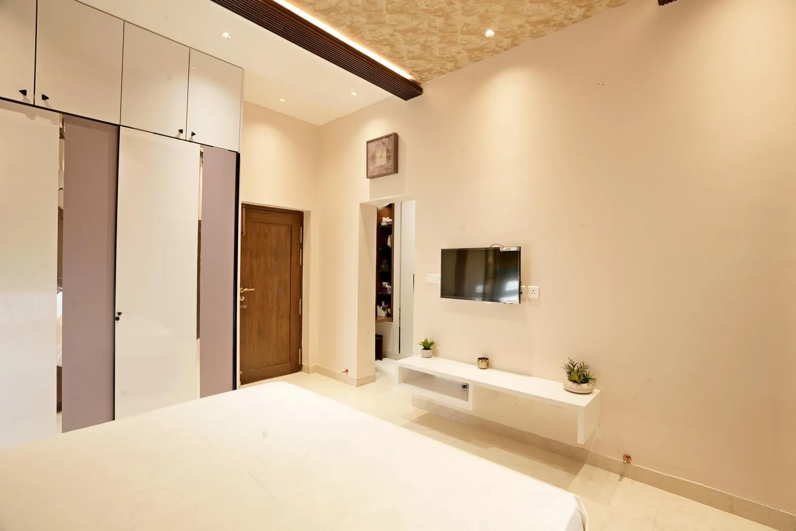 The room includes a built-in wardrobe with sleek finishes and polished flooring. Wooden accents, layered ceiling lighting, and minimal décor create a bright, functional, and balanced modern space.