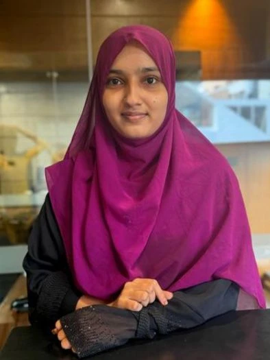 A woman wearing a magenta hijab and a black outfit sitting at a table indoors with a wooden wall and a glass window in the background.