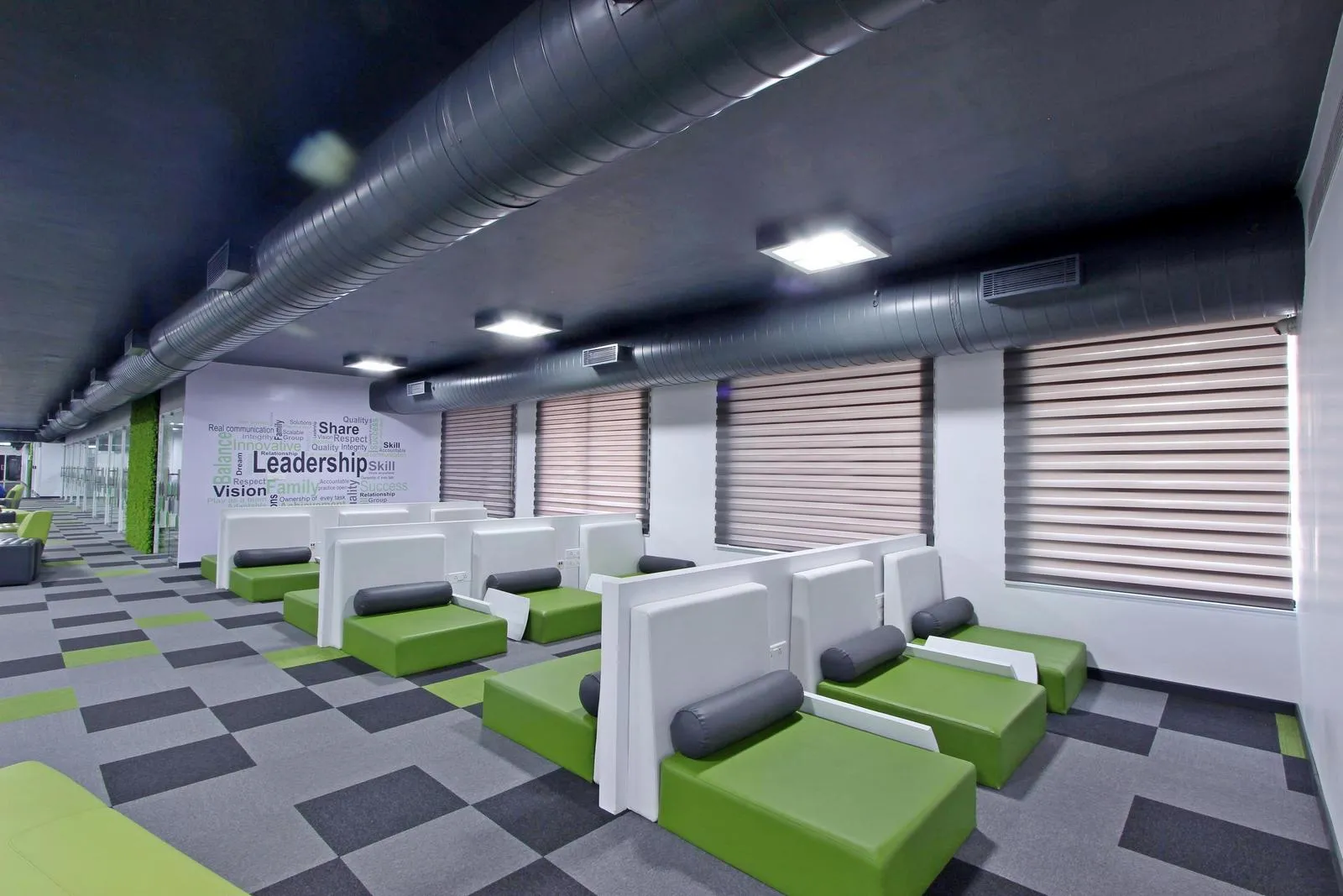 The lounge features modular lime green and white seating pods, geometric carpet patterns, and a motivational typography wall, creating a dynamic, brand-focused workspace. Matte ceilings with exposed ducts, dual-tone blinds, and recessed LED lighting enhance industrial-modern style and visual comfort.