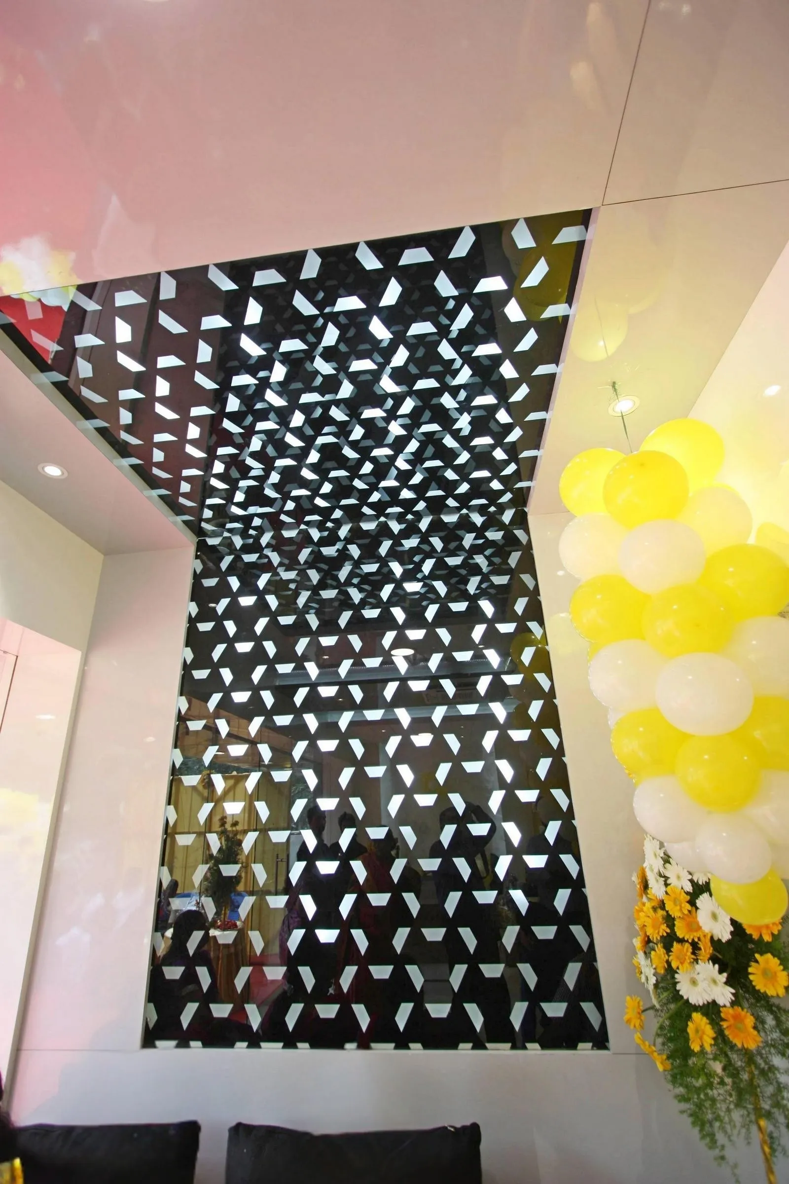 The interior showcases a modern, elegant focal point with a geometric feature wall and ceiling that blend reflective materials and clean finishes. High-gloss panels, mirrored inlays, and subtle lighting enhance depth and sophistication, while seating and floral accents balance comfort with visual drama.