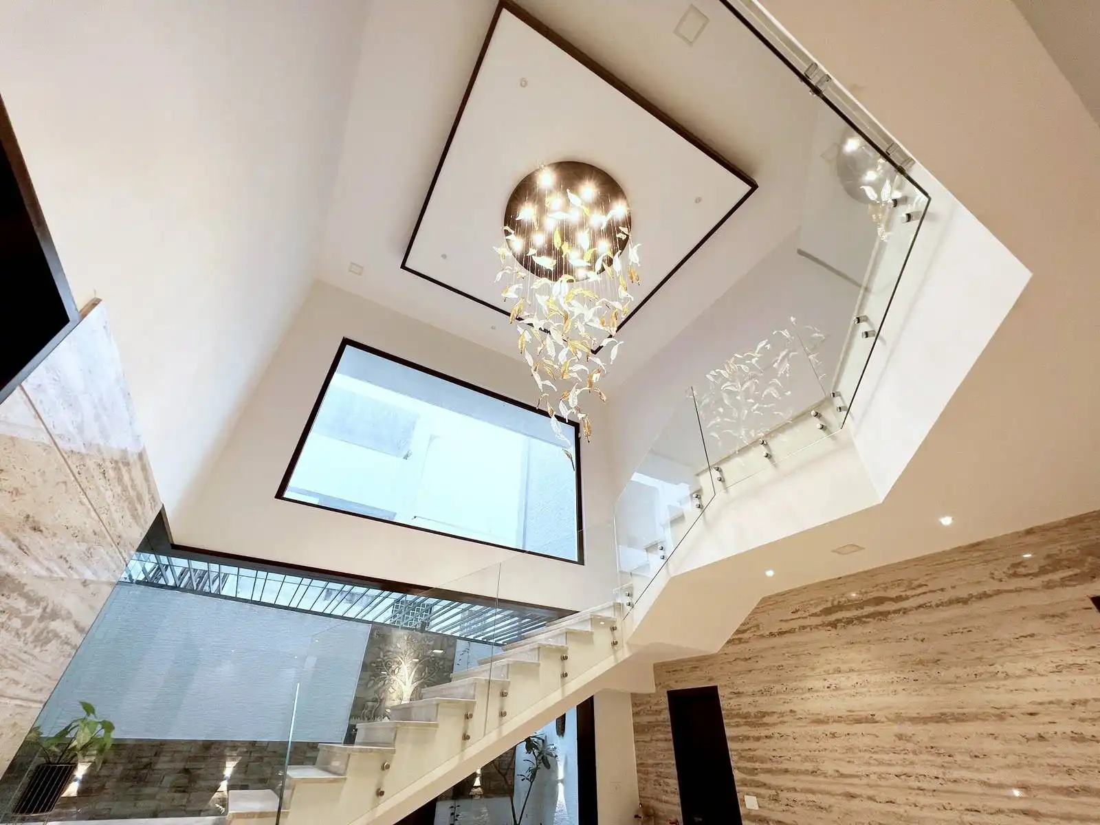 The balustrade is made of clear glass panels with sleek metal handrails, maintaining transparency and safety while maximizing light flow. The surrounding walls feature a textured stone or tile finish, adding warmth and visual interest to the space.
