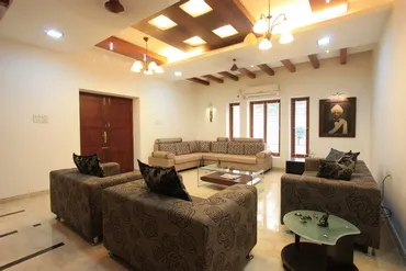 Modern living room with three brown patterned sofas arranged around a wooden center table, featuring ceiling lights, a wooden door, large windows, and a framed portrait on the wall.