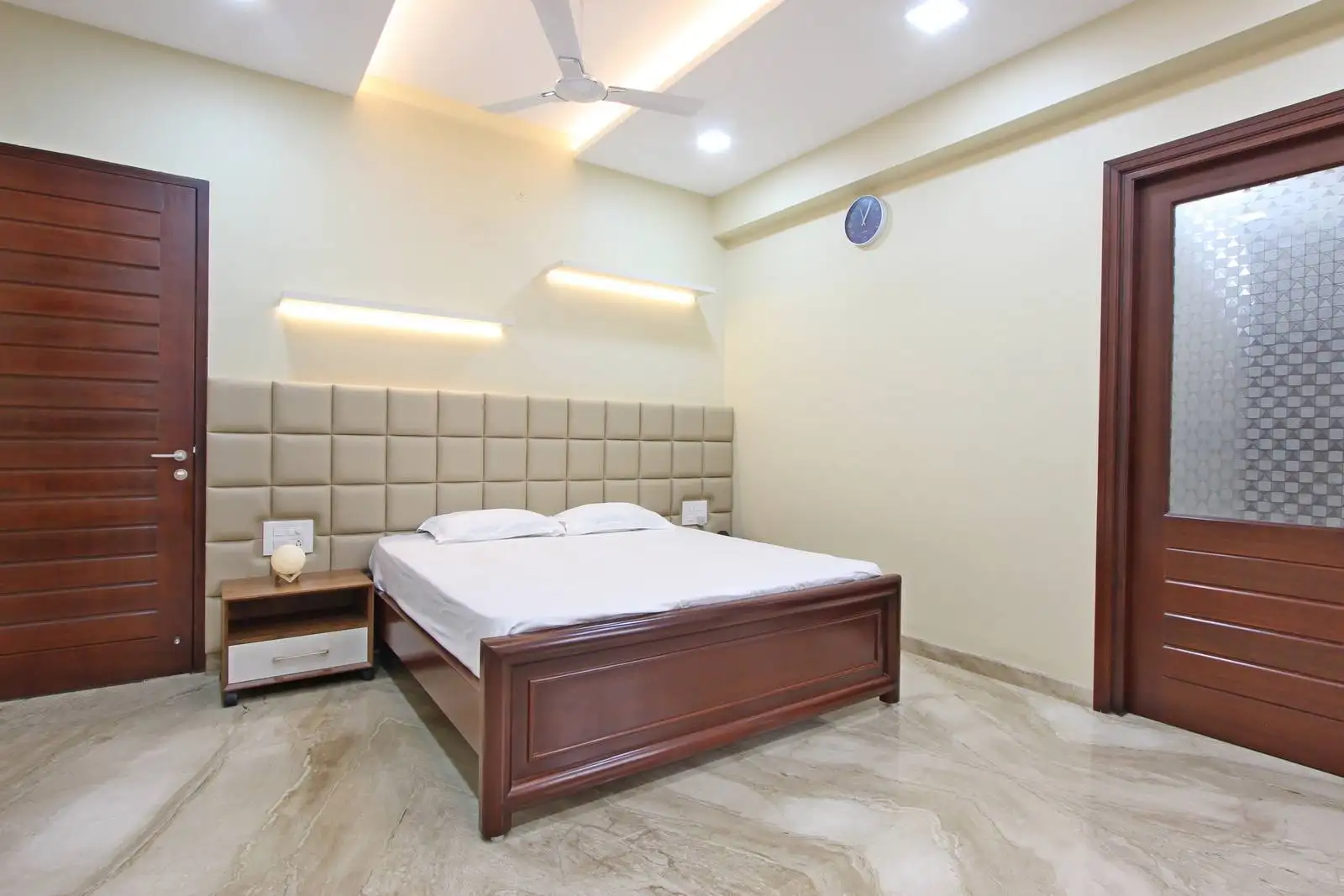 Cozy bedroom featuring a wooden double bed with white bedding, beige padded headboard, marble flooring, and minimal decor for a clean, restful ambiance.