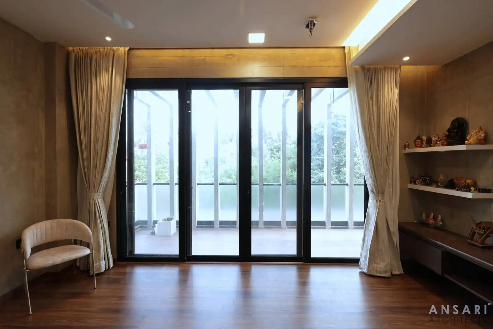 Interior space with large black-framed sliding glass doors opening to a balcony, light beige curtains tied back on either side, and wooden flooring.