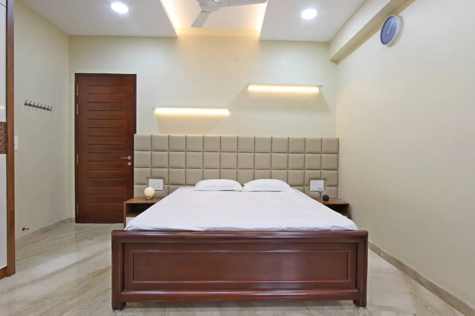 Polished marble flooring, a dark wood bed frame, and a beige upholstered headboard create a warm, elegant bedroom. Layered lighting with recessed and LED accents enhances both ambiance and function.