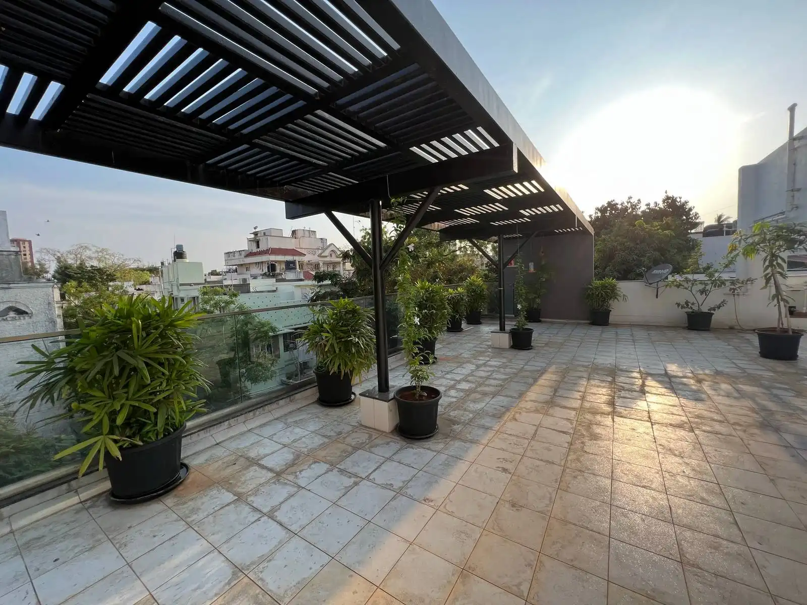 A spacious terrace with light-tiled flooring, sleek black pergola, glass railings, and lush greenery, offering versatile sunlit and shaded outdoor living.