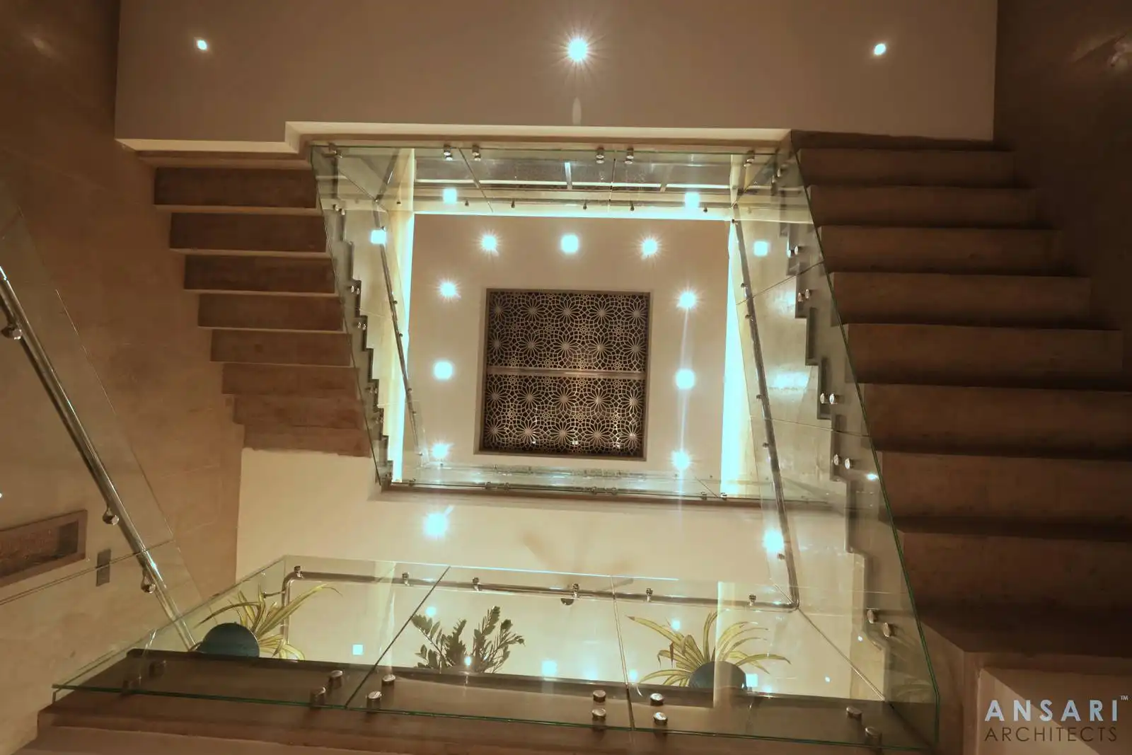 Designed indoor staircase with clear glass railings, recessed lighting, and a decorative ceiling panel, combining transparency and intricate detailing for a striking modern look.