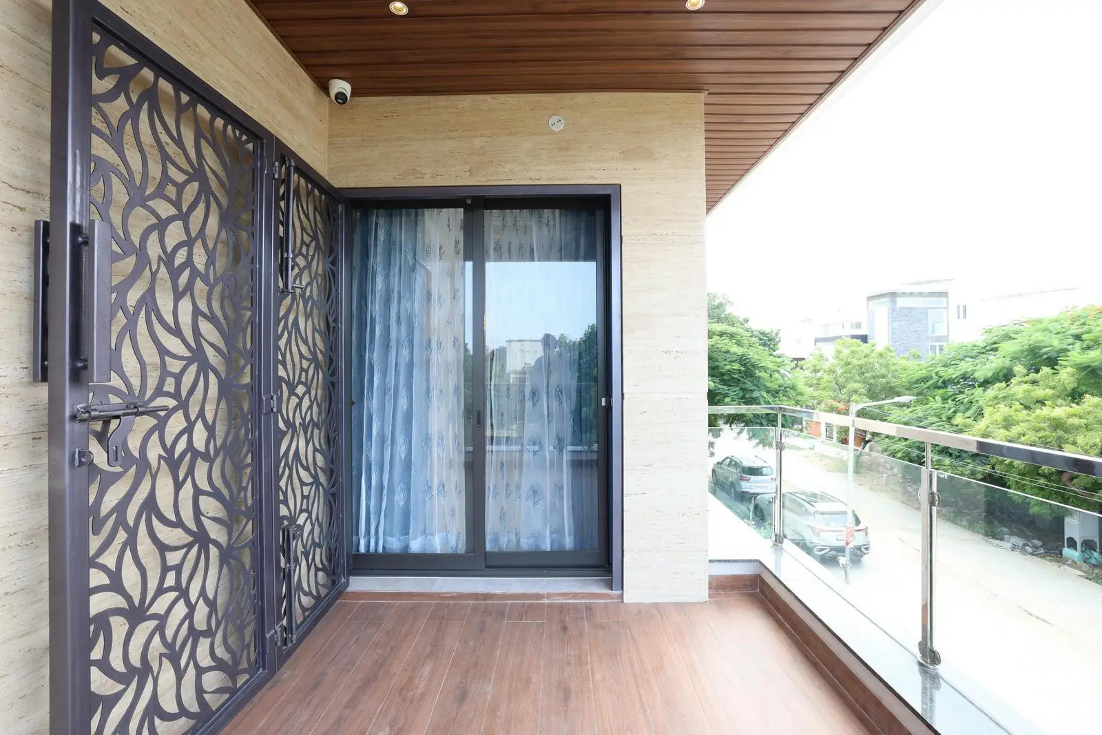 The balcony has wood-finish flooring, glass railings, and beige travertine walls for a warm, modern feel. Sliding doors, recessed lighting, and metal grill details enhance openness and the connection to the outdoors.