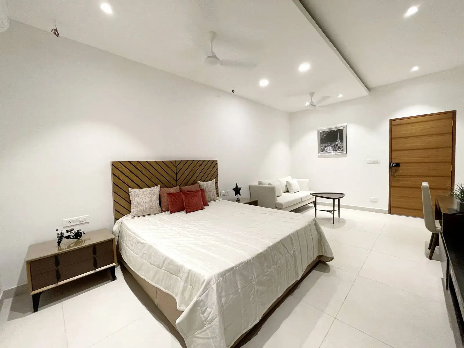 This bedroom features a central wooden headboard, neutral tones, and terracotta accents for a balanced minimalist aesthetic. Integrated lighting, compact seating, and large-format flooring enhance comfort, flow, and spatial openness.