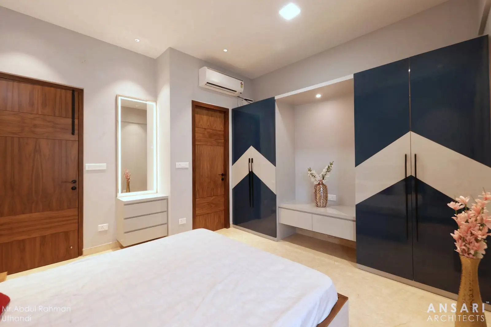 Wardrobes in glossy navy and beige with a chevron pattern add contemporary flair, balanced by warm wooden doors and neutral flooring for a harmonious, inviting bedroom.