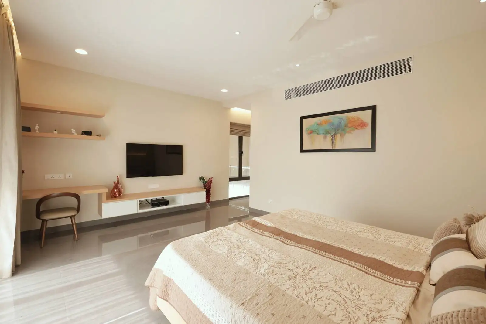 The bed is positioned to one side, allowing for a clear view of the TV and entertainment unit on the opposite wall. A dedicated workspace is created with a floating desk and chair in the corner, making the room multifunctional.