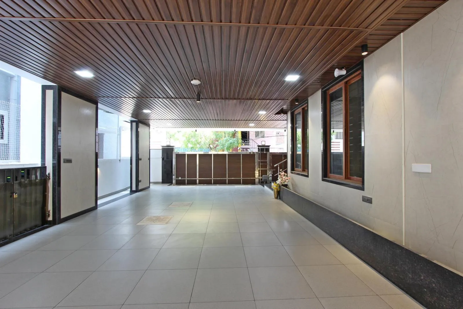 The car parking is a covered, semi-open space with a rectangular layout for easy vehicle movement. Light grey vitrified floors, wooden ceiling slats, and beige walls create a functional, elegant space with natural light and ventilation.