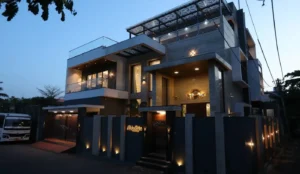 Modern multi-level house at dusk with exterior lights illuminating the entrance and perimeter, featuring large windows, balconies with glass railings, and a decorative sun-shaped wall light near the front door.