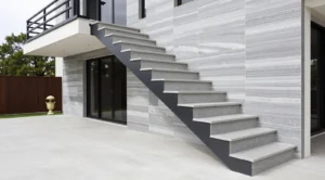 Modern outdoor staircase with grey stone steps and black railing, attached to a building with grey striped stone walls and large sliding glass doors, with a tiled floor and greenery in the background.