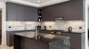 Modern kitchen with dark gray cabinets, black granite countertops, a central island with a sink and faucet, marble backsplash, built-in stainless steel oven and gas stove, and under-cabinet lighting.