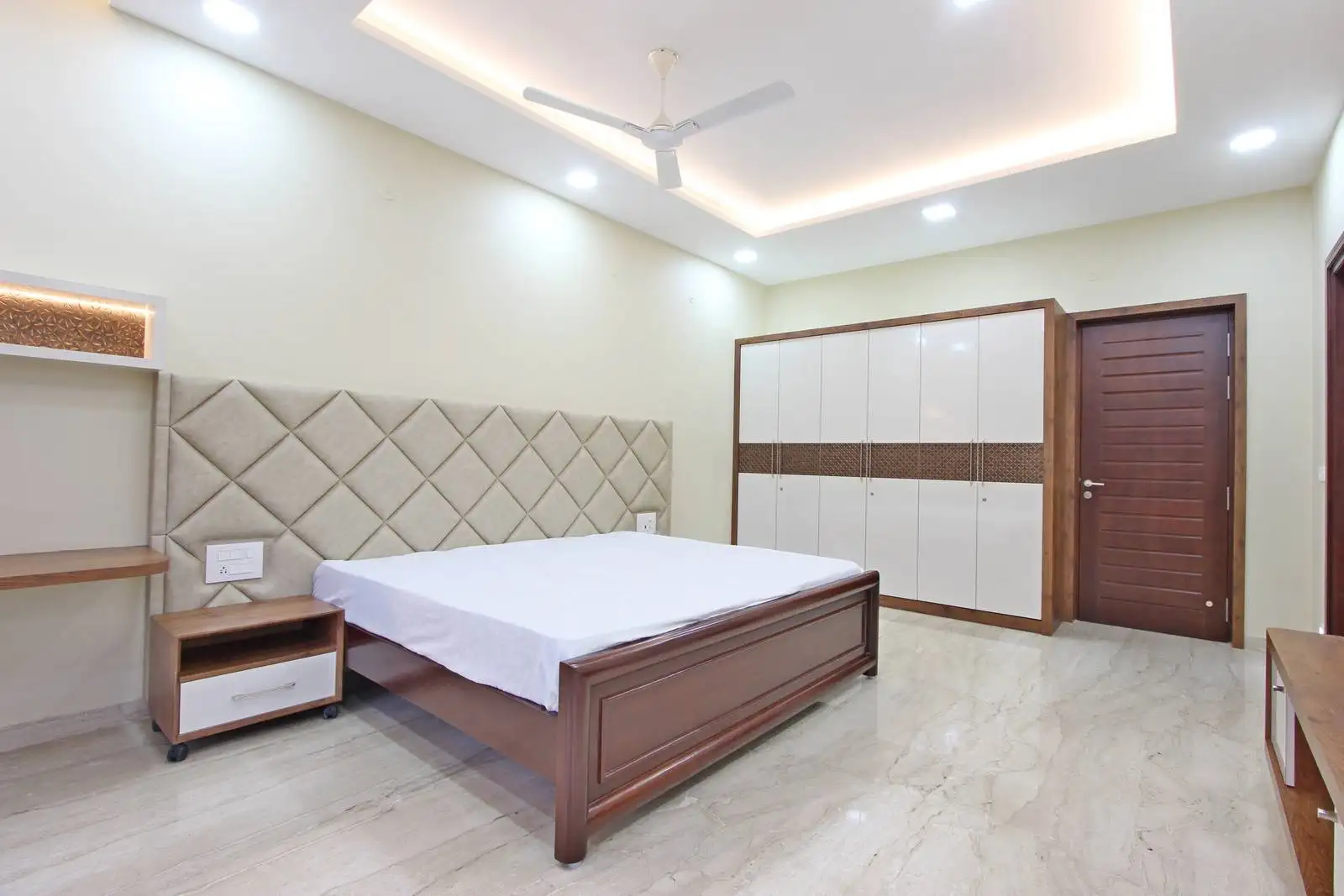 A bedroom with a diamond-patterned upholstered headboard, ambient recessed and cove lighting, and focused lighting over the desk, blending comfort with functional design.