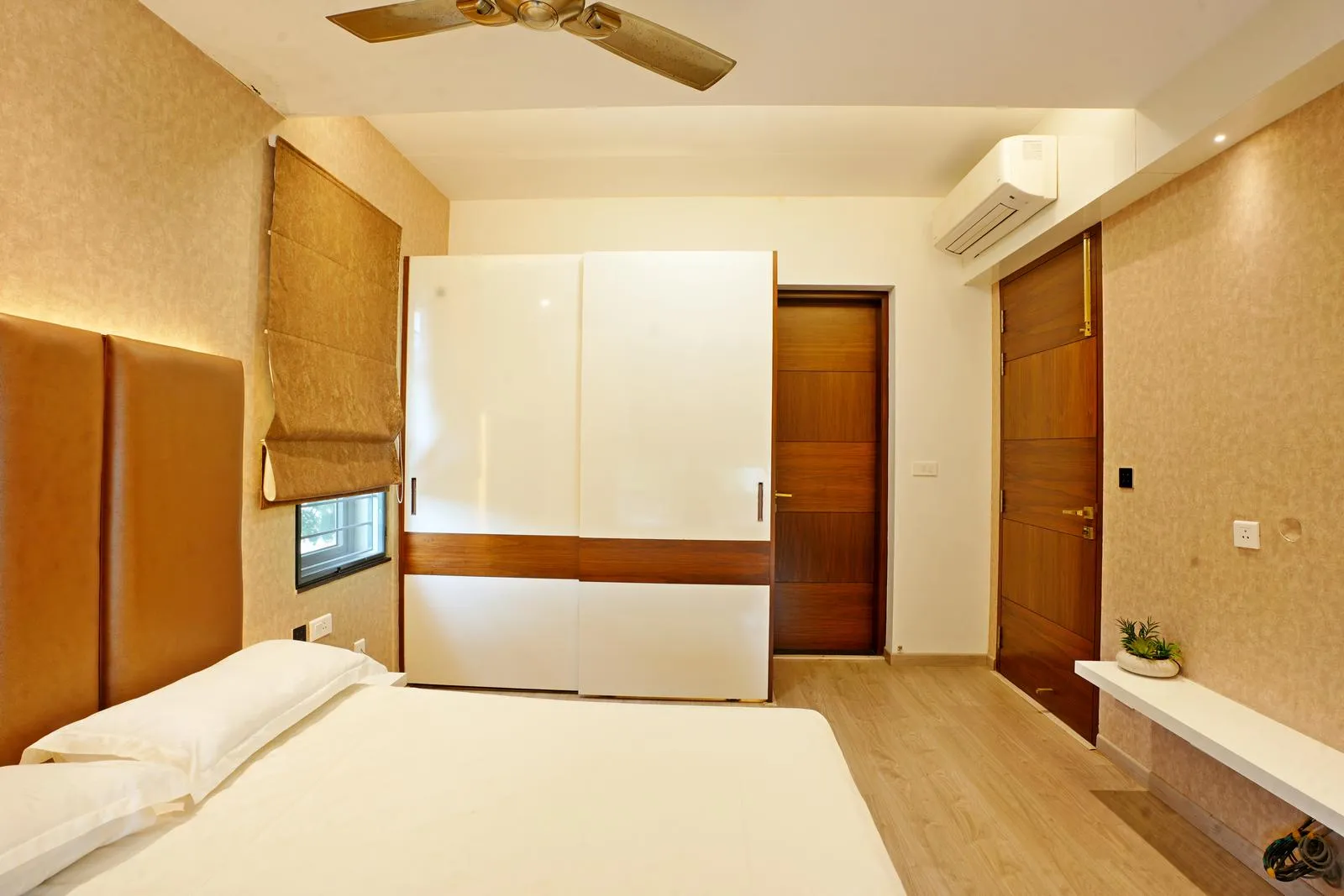 Warm beige walls, light oak flooring, and a wardrobe combining white laminate with wood veneer. A tan leatherette headboard and wooden doors add texture and continuity, creating a cozy, modern space.