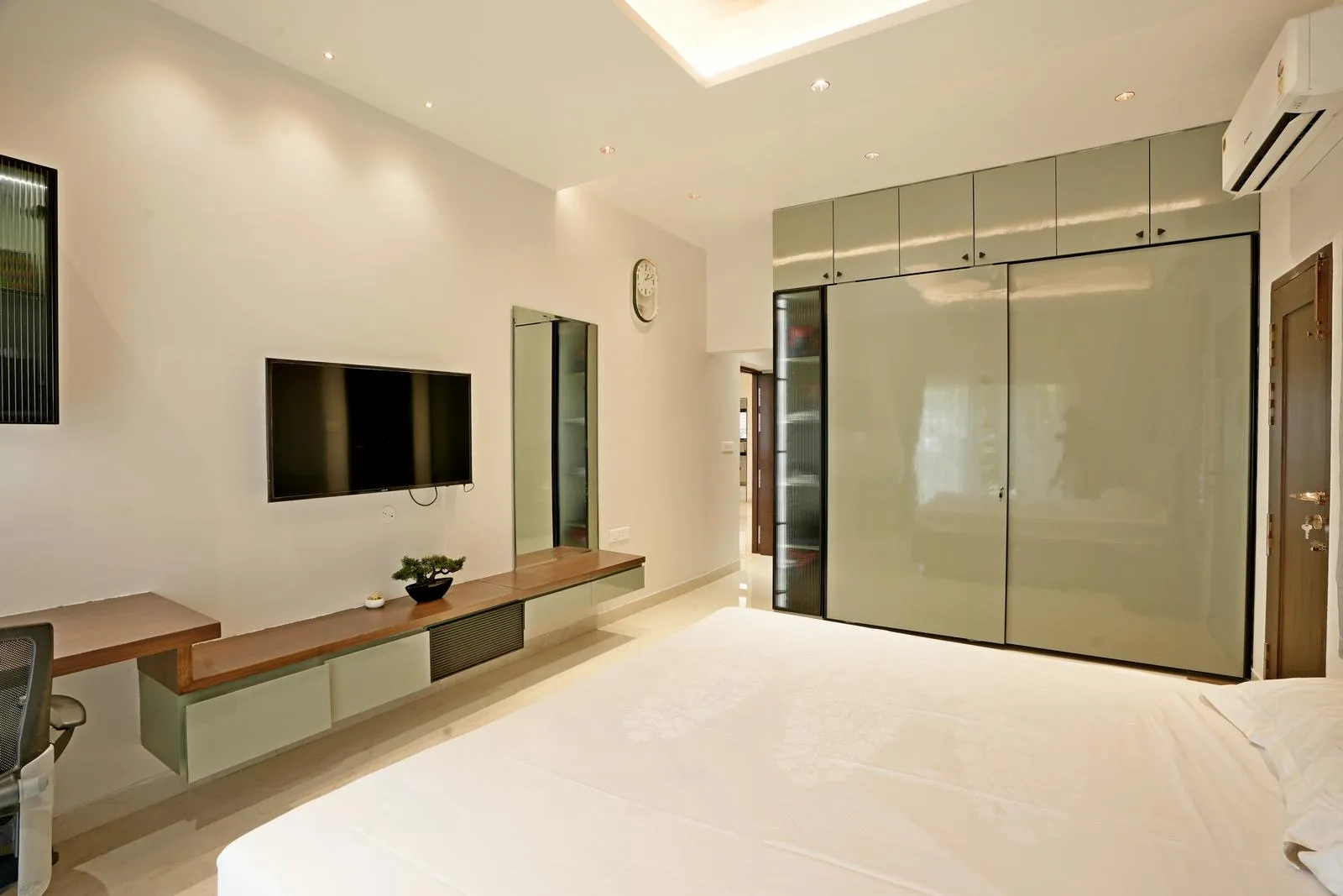 The bedroom features cream vitrified flooring and sleek grey wardrobes. Floating TV units, mirror panels, and layered ceiling lighting create a bright, airy, and minimalist space with functional elegance.