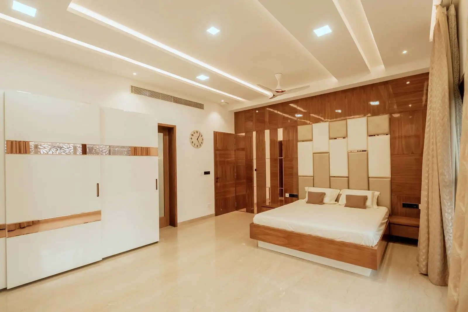 Bedroom features a high-gloss wood and upholstered accent wall paired with a sleek white wardrobe enhanced by wooden and metallic detailing for a refined modern look.