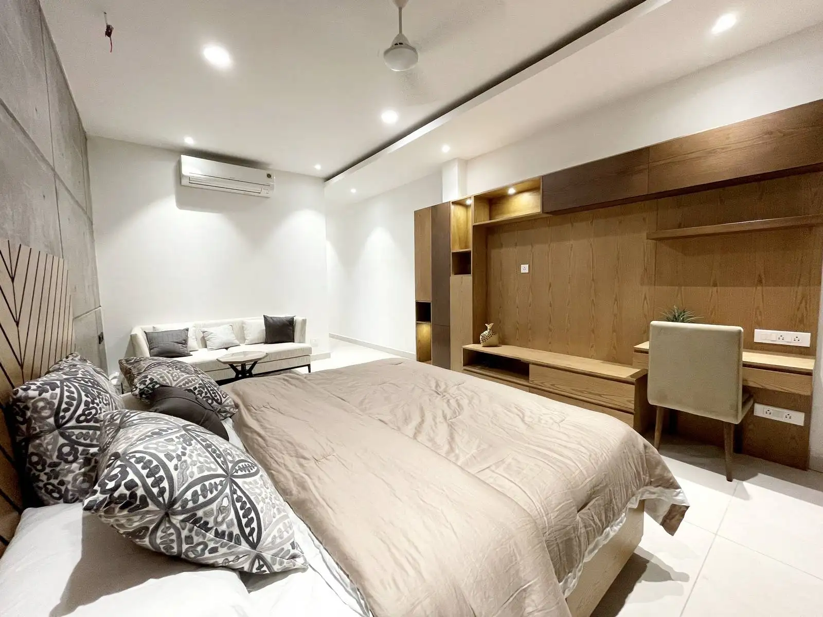 The bedroom features exposed concrete, warm timber, and neutral fabrics in a clean, linear layout with an integrated study and seating area. Strategic lighting and precise joinery enhance comfort, clarity, and refined architectural detail.