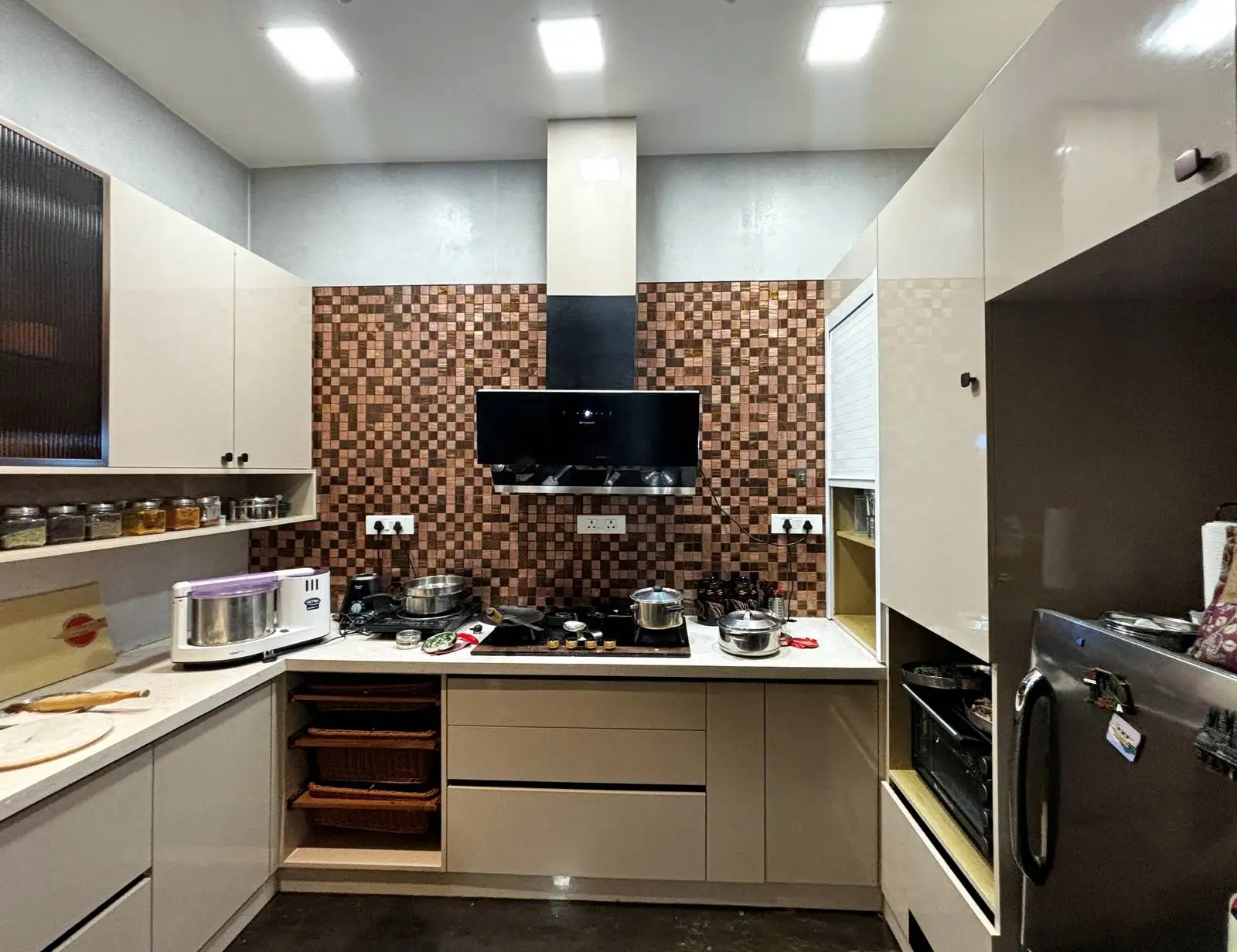 The cabinetry features a sleek, glossy finish in neutral tones, contributing to a clean and modern aesthetic. The backsplash is a standout feature, using a mosaic of brown tiles that adds warmth, texture, and visual interest to the space.