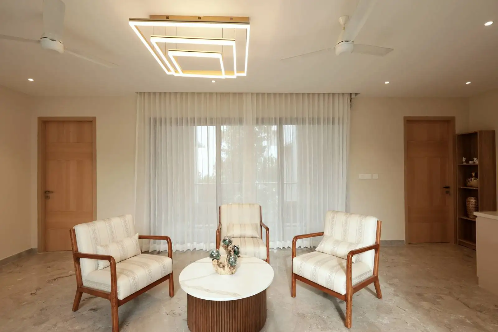 The family room showcases a serene ambiance with cream-colored armchairs, a marble coffee table, sheer white curtains, and warm wooden accents, all under soft geometric pendant lighting for a refined, contemporary feel.
