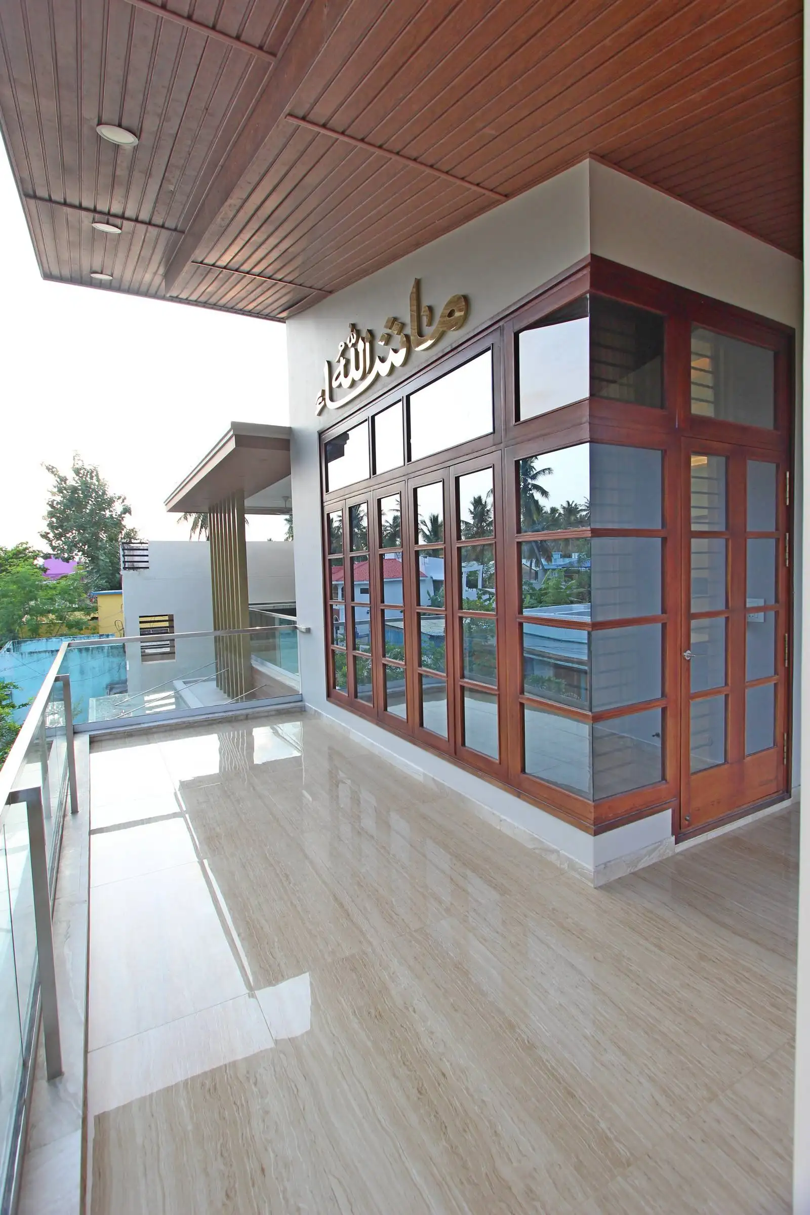 The balcony is spacious and elongated, running alongside a room with large glass windows and a wooden-framed door, allowing for seamless indoor-outdoor connectivity.
