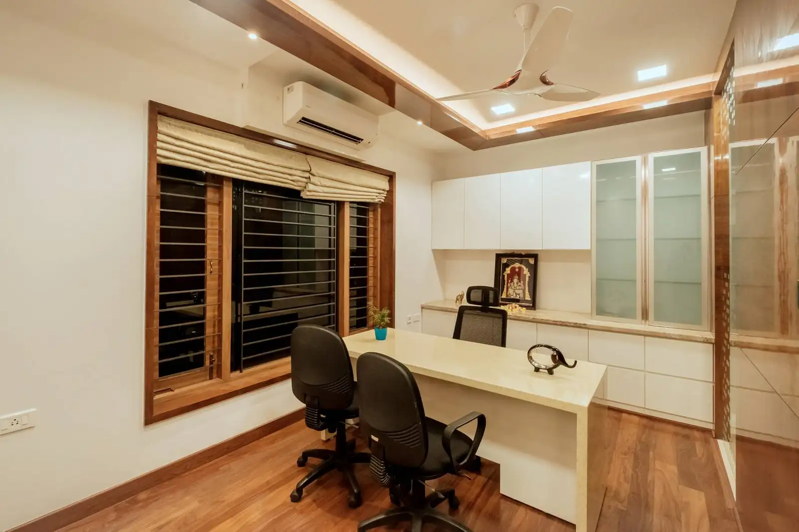 A sophisticated office featuring a central desk, rich wood flooring, built-in cabinetry, layered lighting, and a large window with blinds for a professional and comfortable workspace.