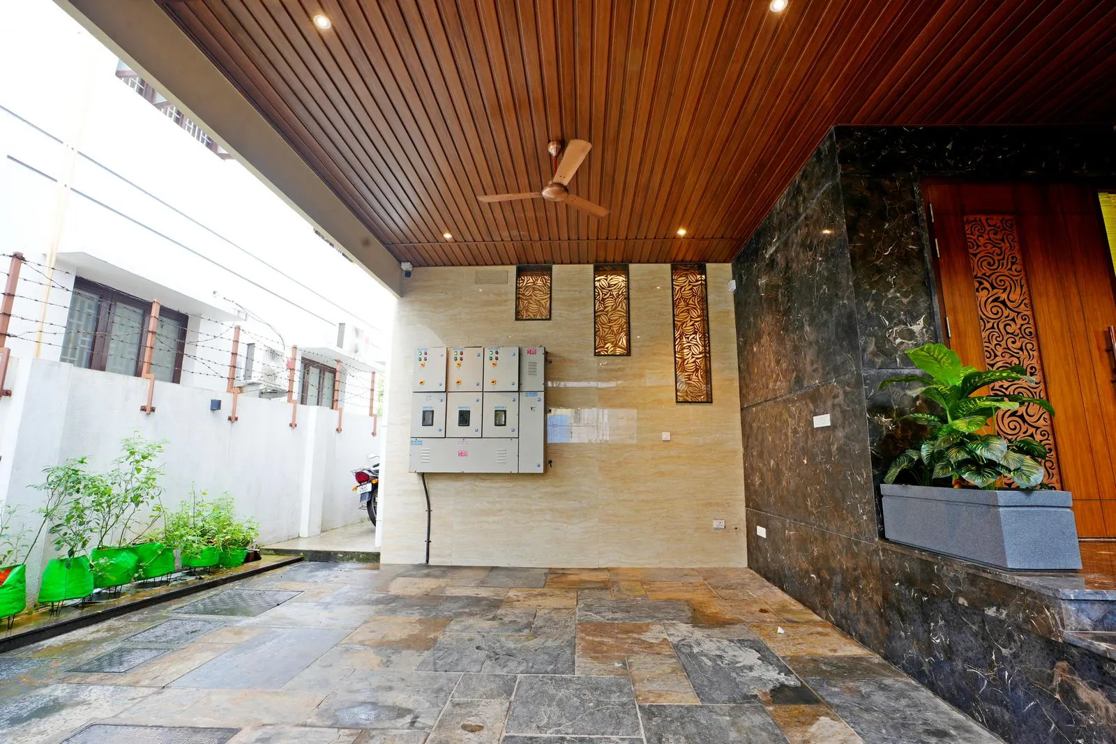 The 18'0" × 20'0" sq. ft car porch functions as a seamless transition from driveway to entrance, offering efficient circulation and openness. Natural stone flooring, wooden ceiling slats, integrated lighting, and greenery create a durable, elegant, and harmonious space.