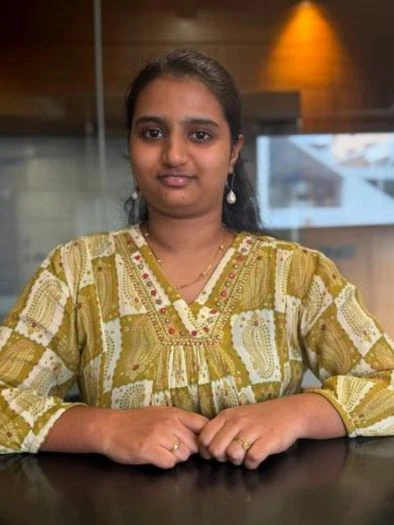Portrait of a woman wearing a yellow patterned top sitting with her hands folded on a dark surface in an indoor setting with a warm light in the background