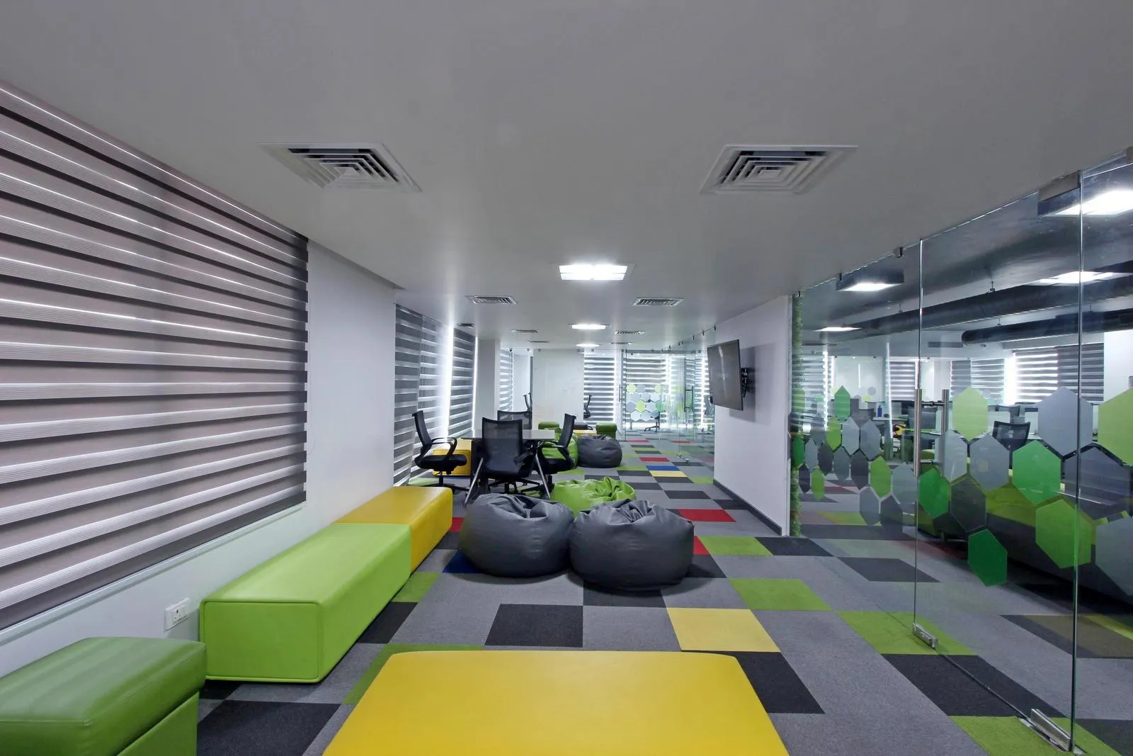 The informal lounge features modular lime green and yellow seating, charcoal bean bags, and patterned carpet, creating a vibrant, collaborative, and flexible workspace. Frosted glass partitions, recessed LED lighting, and integrated AC enhance comfort, openness, and functionality.