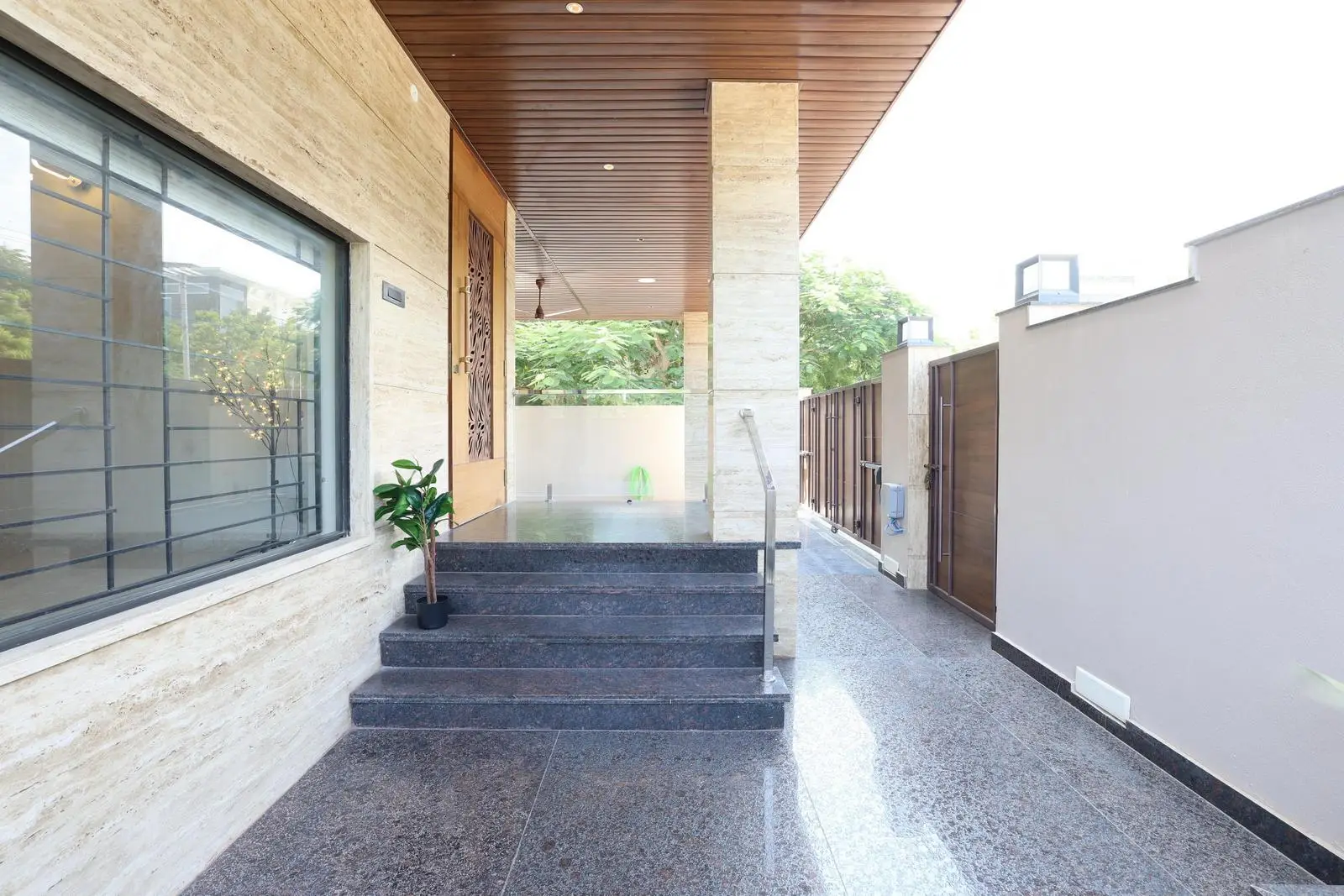 The verandah guides visitors from gate to entrance with an elevated platform. Dark granite floors, travertine walls, and wooden ceilings create a warm, elegant palette. Metal accents, window grills, and planters enhance modern aesthetics while ensuring light, safety, and style.